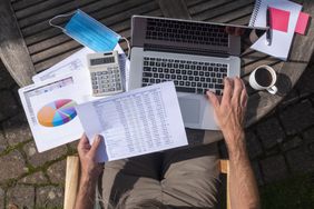 A person seated outside, picture taken from above looking down, as they review a list of stocks with one hand on a laptop.