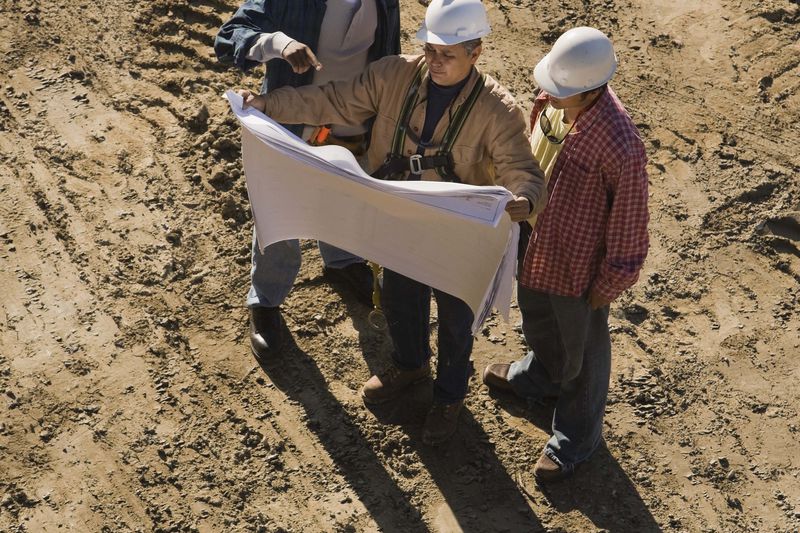 An engineer, an architect, and an earthmoving equipment operator look at a set of site plans while standing in the center of a construction site.