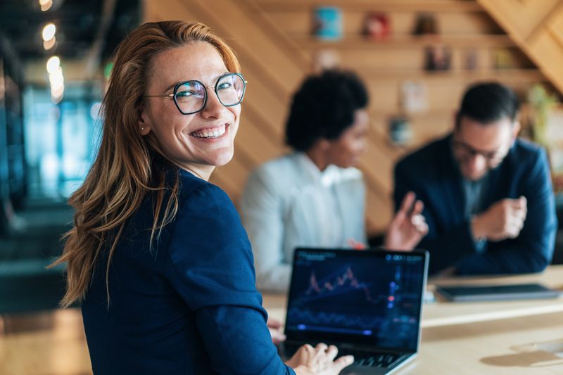 A woman smiling and working on a laptop, with two people discussing in the background