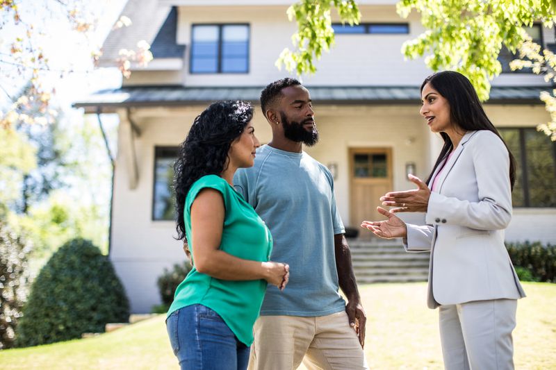 A couple discuss upcoming escrow procedures with their real estate agent in the yard front of the two-story house they may buy.