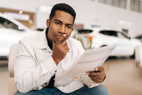 A young man seated at a car dealership, holding a document and looking thoughtful