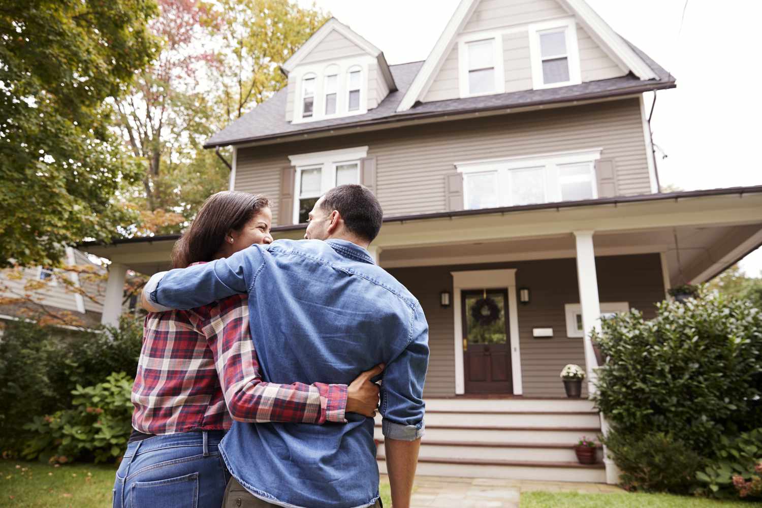 A man and a woman stand out front of a beautiful tan home with a large porch with their arms around each other