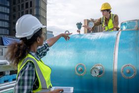 Two workers in safety gear interacting near a large industrial tank, one pointing upward and the other standing on the tank