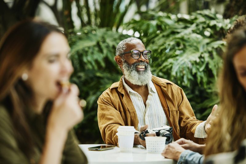 Smiling man with gray hair and a beard sitting in an outdoor cafe holding a camera.