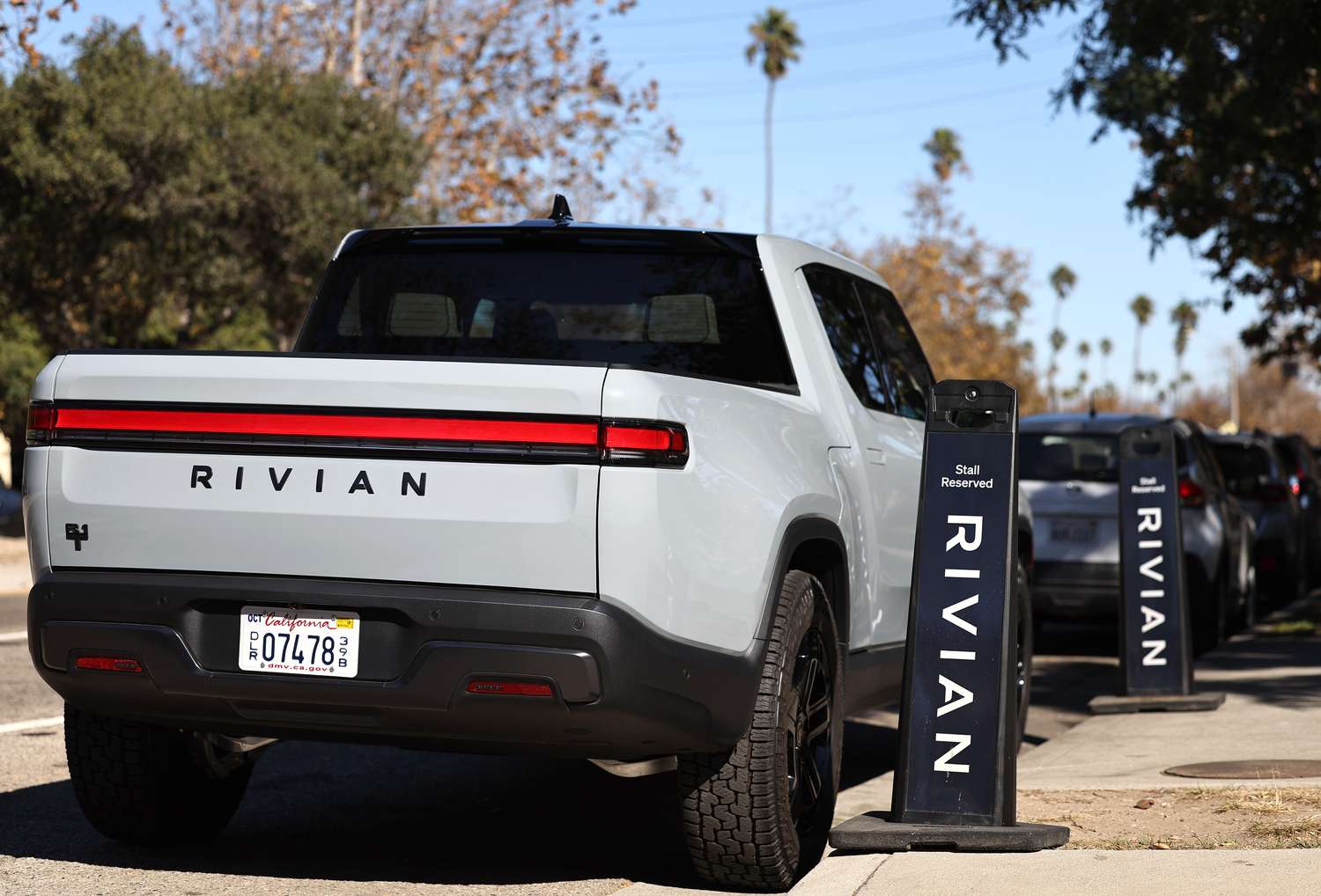 A gray Rivian truck is seen parked next to signs on a sidewalk with the Rivian logo.
