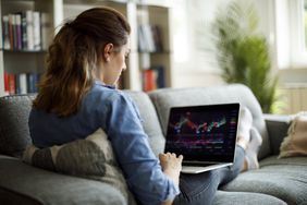 A woman with her back to the camera sits on a couch and looks at a laptop screen depicting stock charts.