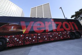 The Verizon logo is seen on the side of a bus with buildings and a blue sky in the backrgound.