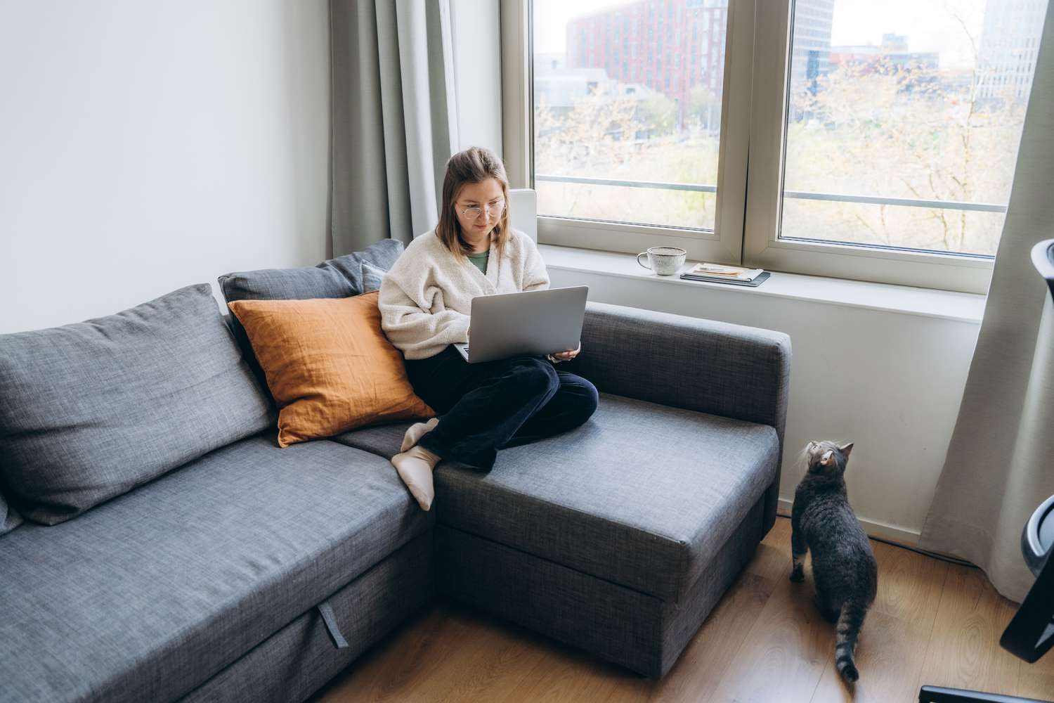 A woman sits on her couch near her window using a laptop while her cat stands next to the couch
