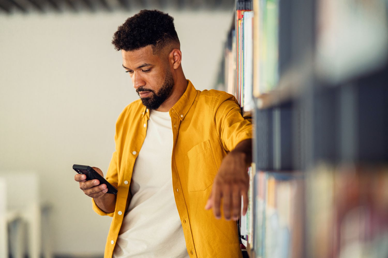 A man standing by bookshelf, text messaging.