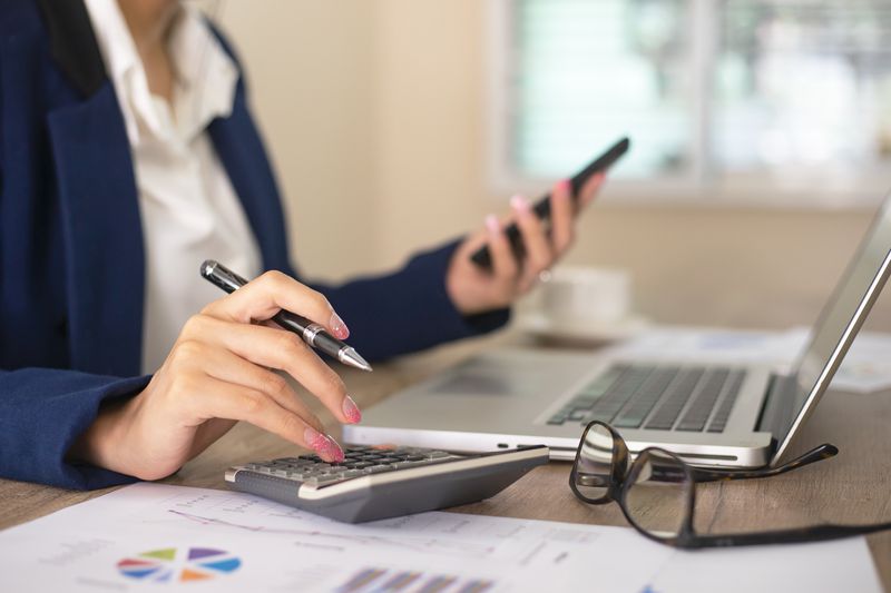 Businesswoman working in office typing on calculator and holding phone in hand with computer and financial sheets on desk