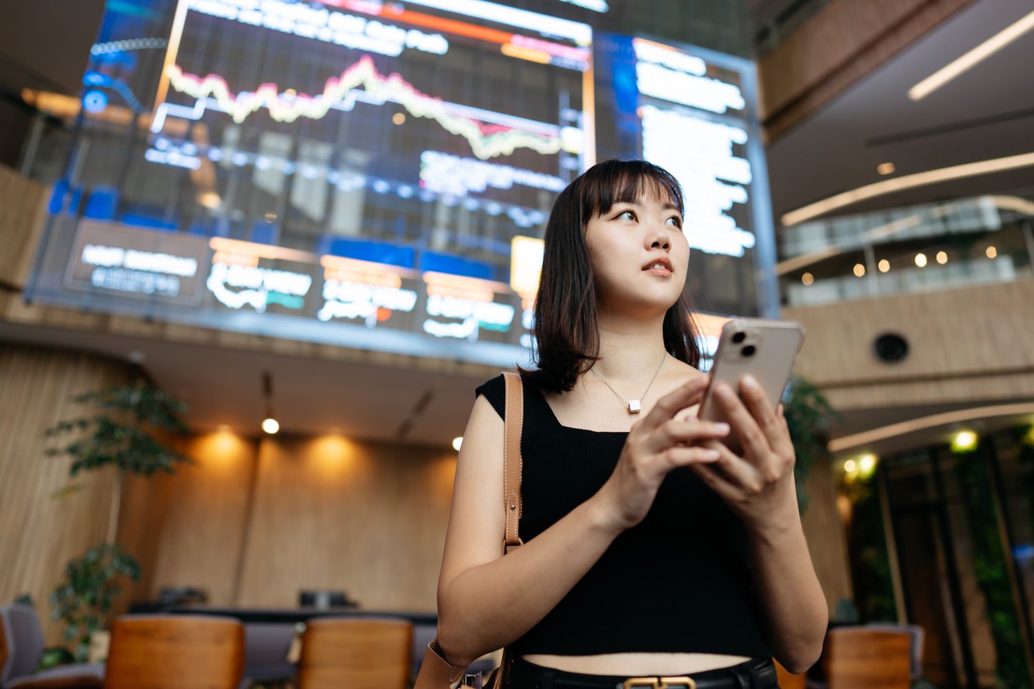Young woman sanding with her back to a large screen showing market data and charts.