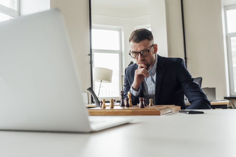 White business man in an office facing a computer with a chess board in front of him. 