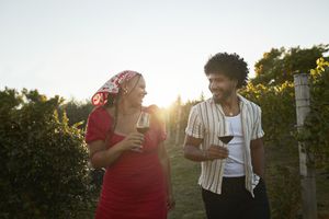 Two people holding wine glasses walking through a vineyard, smiling and enjoying the setting
