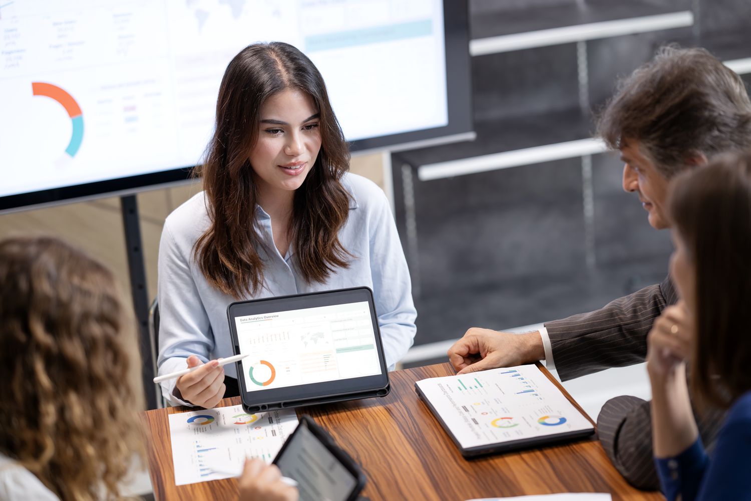 An employee explains a standard budget strategy to other team members using a computer, a large screen, and paper copies of the plan.