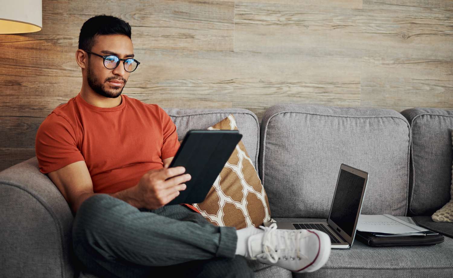 A person sitting on a couch holding a tablet, with a laptop and notebook nearby