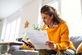 A woman with brown hair and a yellow sweater sits on a couch looking at a document and typing on her cell phone