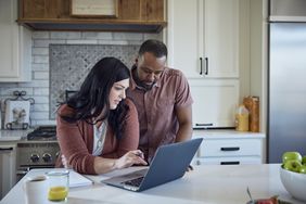 A man and a woman stand in their kitchen looking at a laptop on the kitchen island.