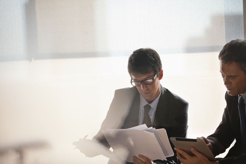 Two business executives look concerned as they shuffle paperwork and use a pad to review information contained on a business report.