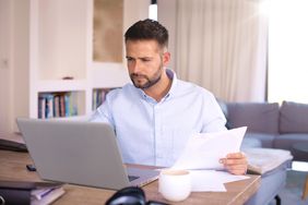 Man in his 30s sitting at his home desk and looking at bank statements and a laptop 
