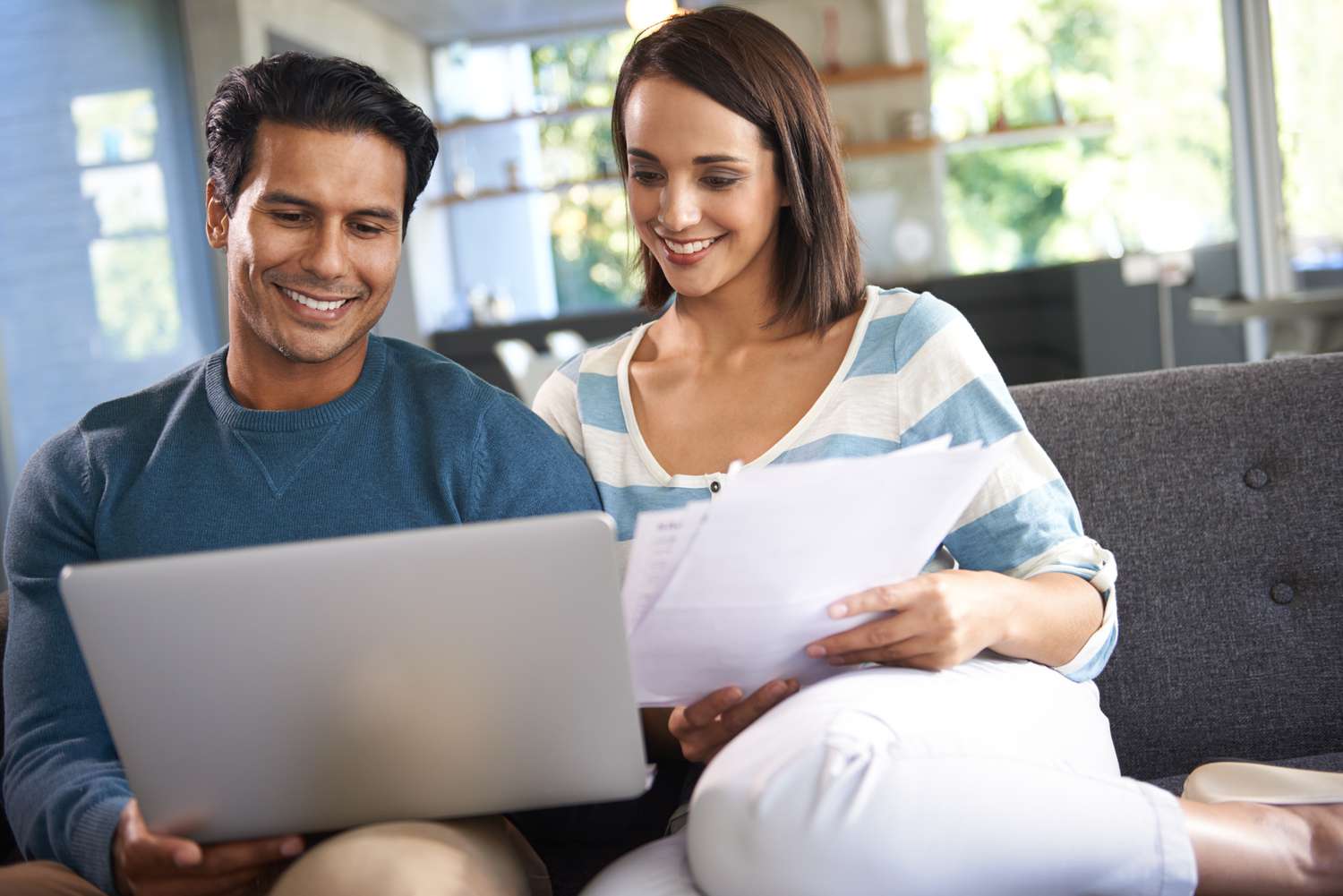 Young couple sitting on their sofa and looking happily together at a laptop and documents