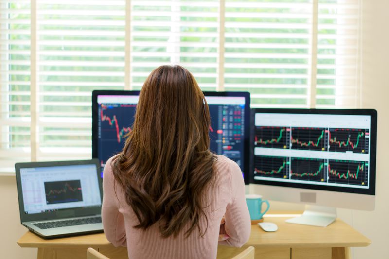 Woman trader at work in home office looking at charts on several monitors