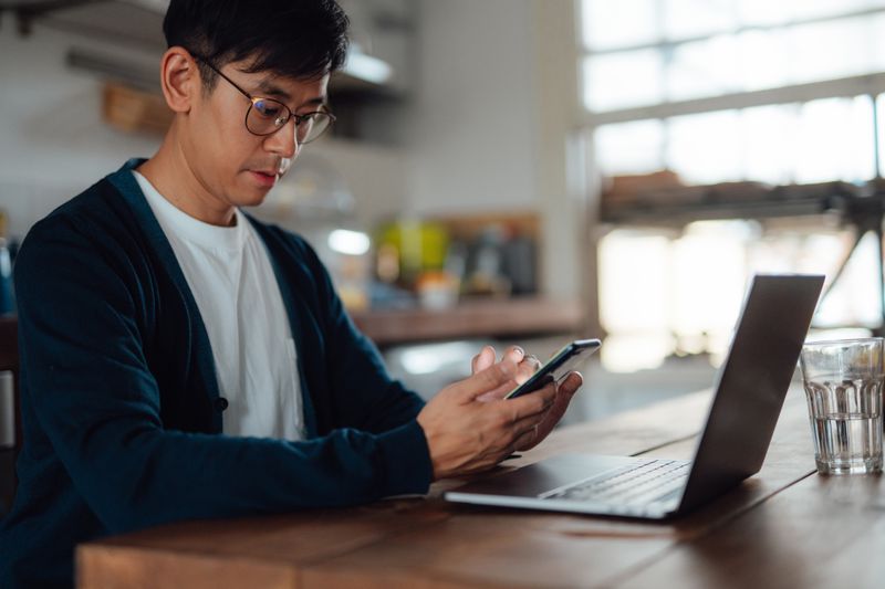 Young Asian man studying phone and laptop at his kitchen table