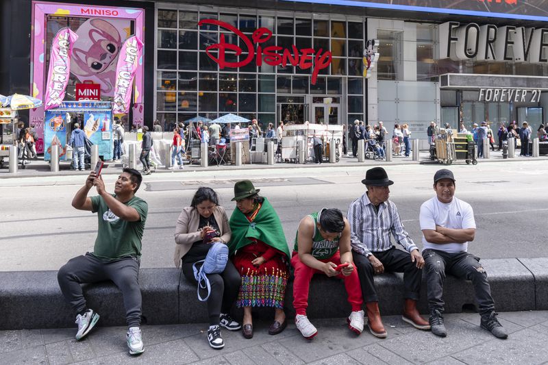 People sitting on a stone ledge in a city setting with a Disney store in the background, holding phones or in conversation