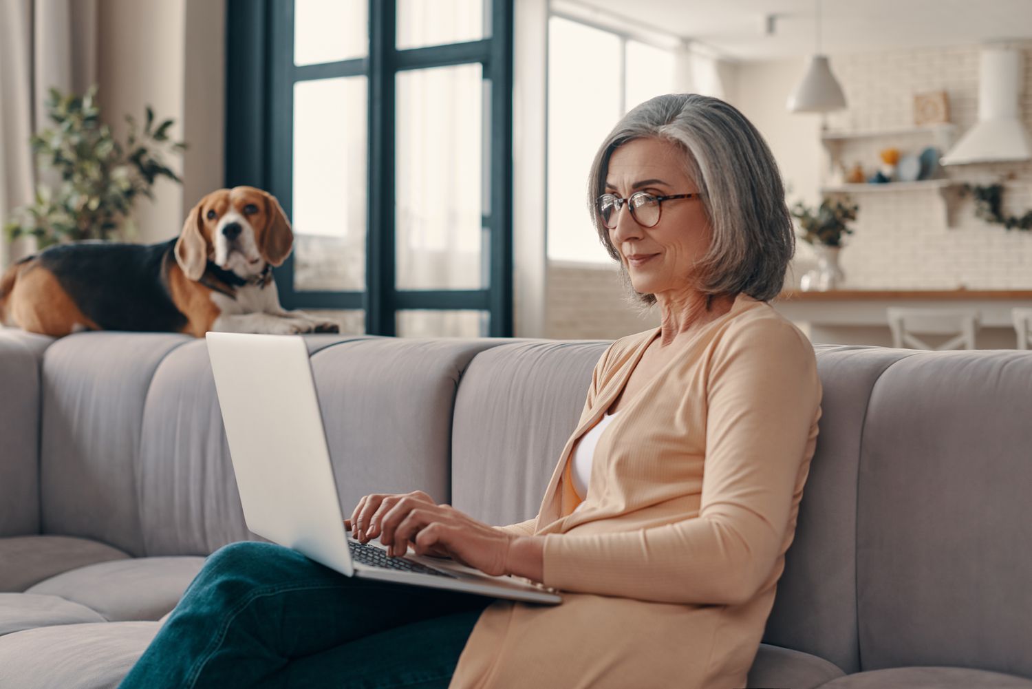 An older woman sits on a couch typing on a laptop while her dog lounges on the couch next to her