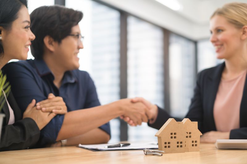 a couple shaking hands with a woman with a model house in front