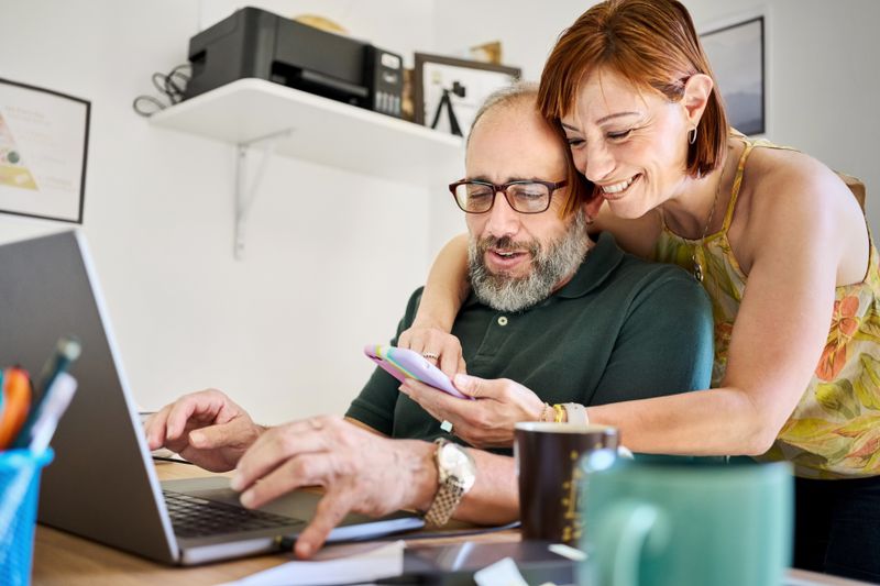 Smiling woman showing her husband something on her phone while he's working on his laptop