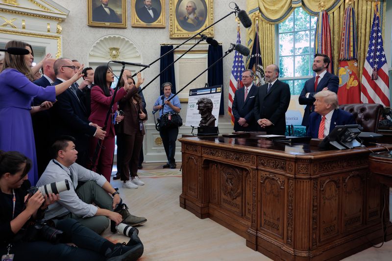 A gaggle of reporters with microphones and cameras stand to left, asking questions of President Donald Trump, who sits behind a desk in the Oval Office. Other administration officials stand behind him, next to a poster explaining a trade agreement between the U.S. and the U.K.