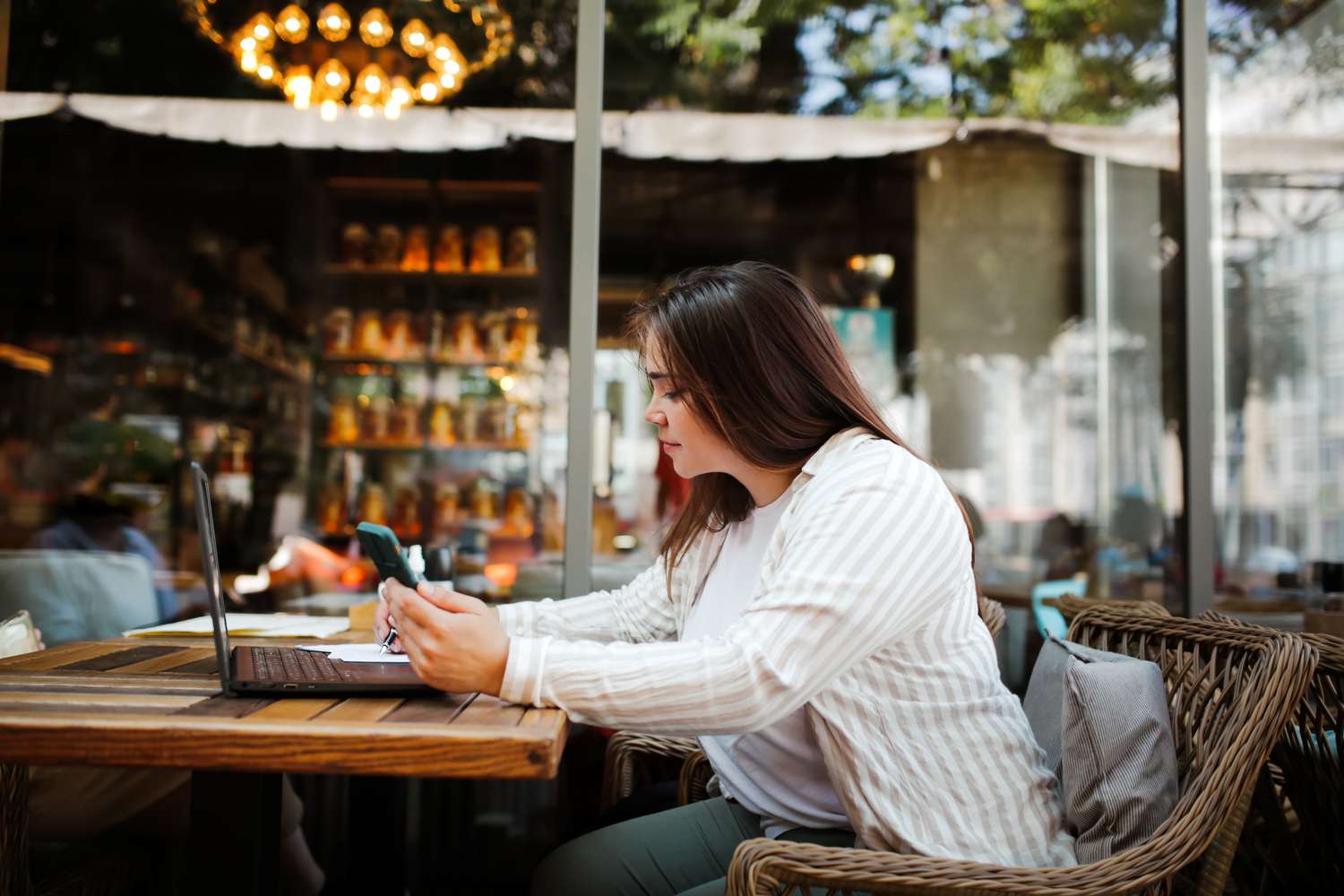A woman works on a laptop and smartphone while sitting at a table in a street cafe. 