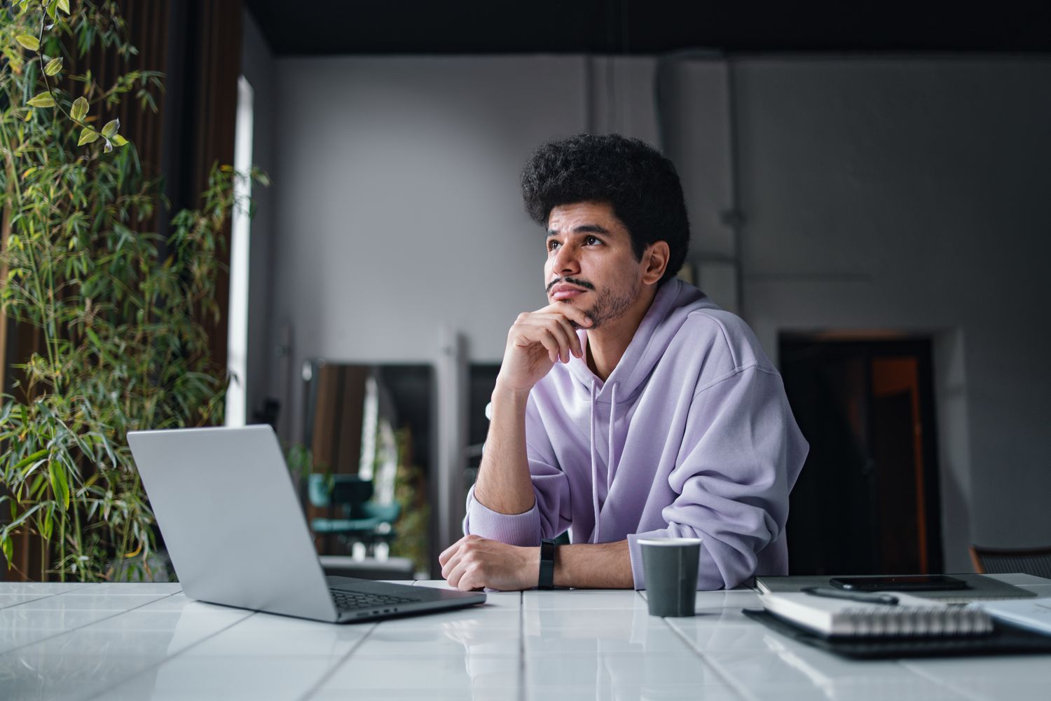 A man sits with a laptop at a table, looking into the distance and pondering.