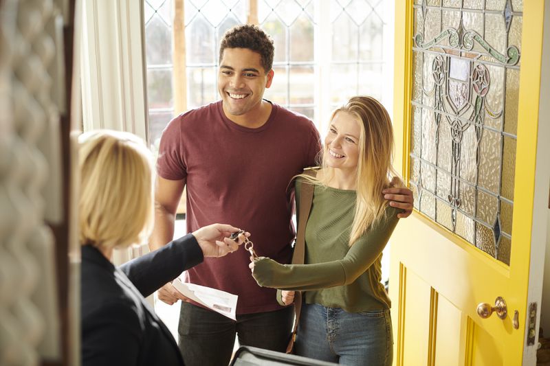 A smiling couple stands at the doorway of their new home, receiving a set of keys from a real estate agent. The bright yellow door and stained-glass window add warmth to the welcoming scene.