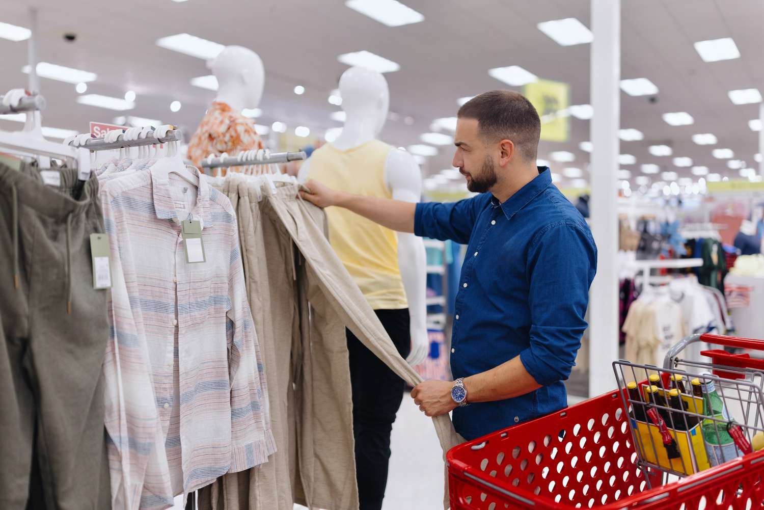 A man in a blue shirt stands next to a red shopping card holding a pair of tan pants in his hands looking at the price