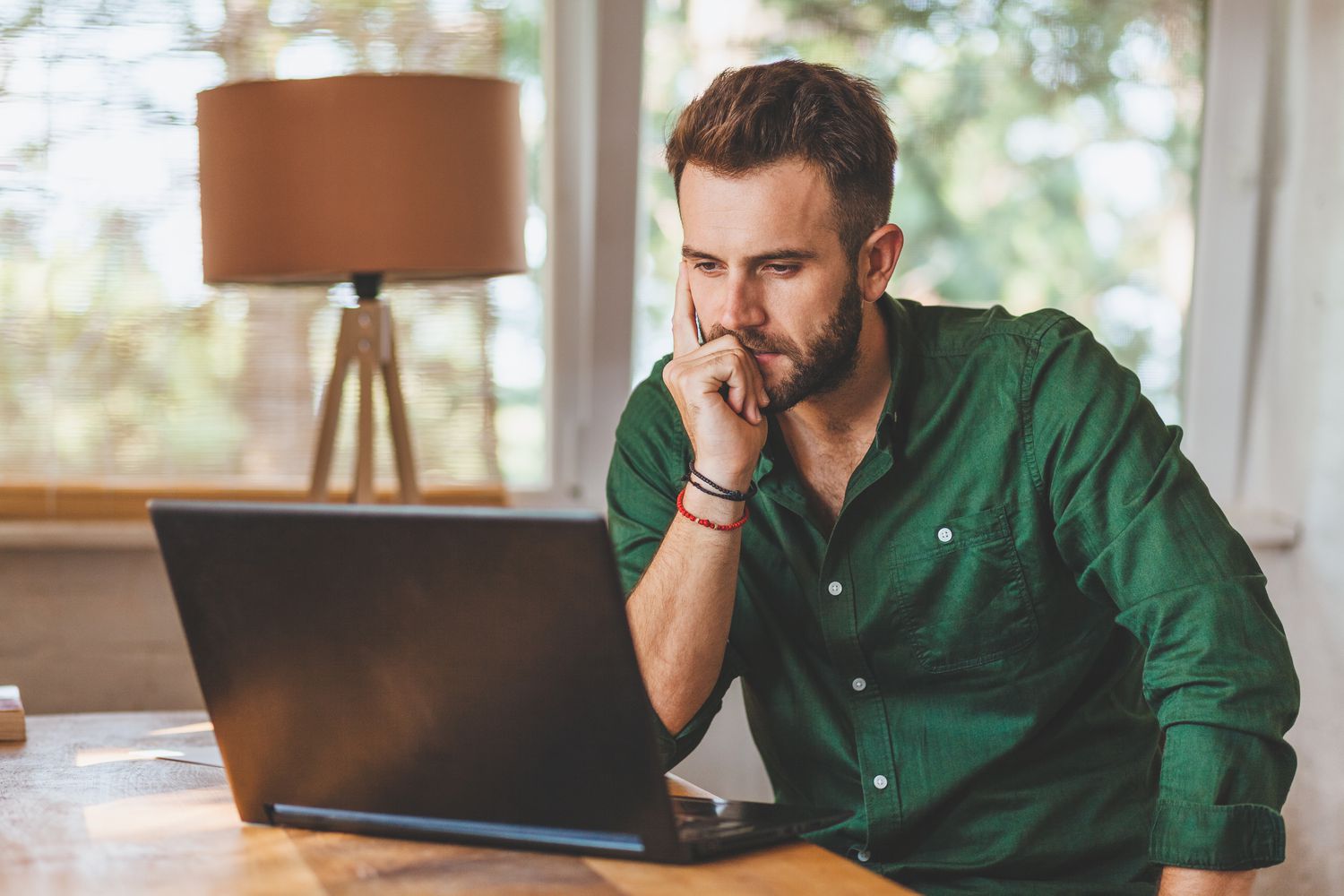 Young man sitting at his dining room table and looking intently at a laptop screen.