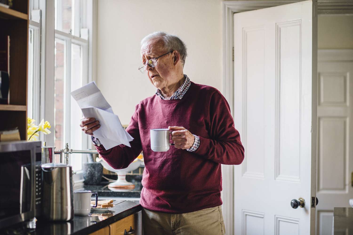 Concerned senior man with paperwork in one hand and a mug of coffee in the other