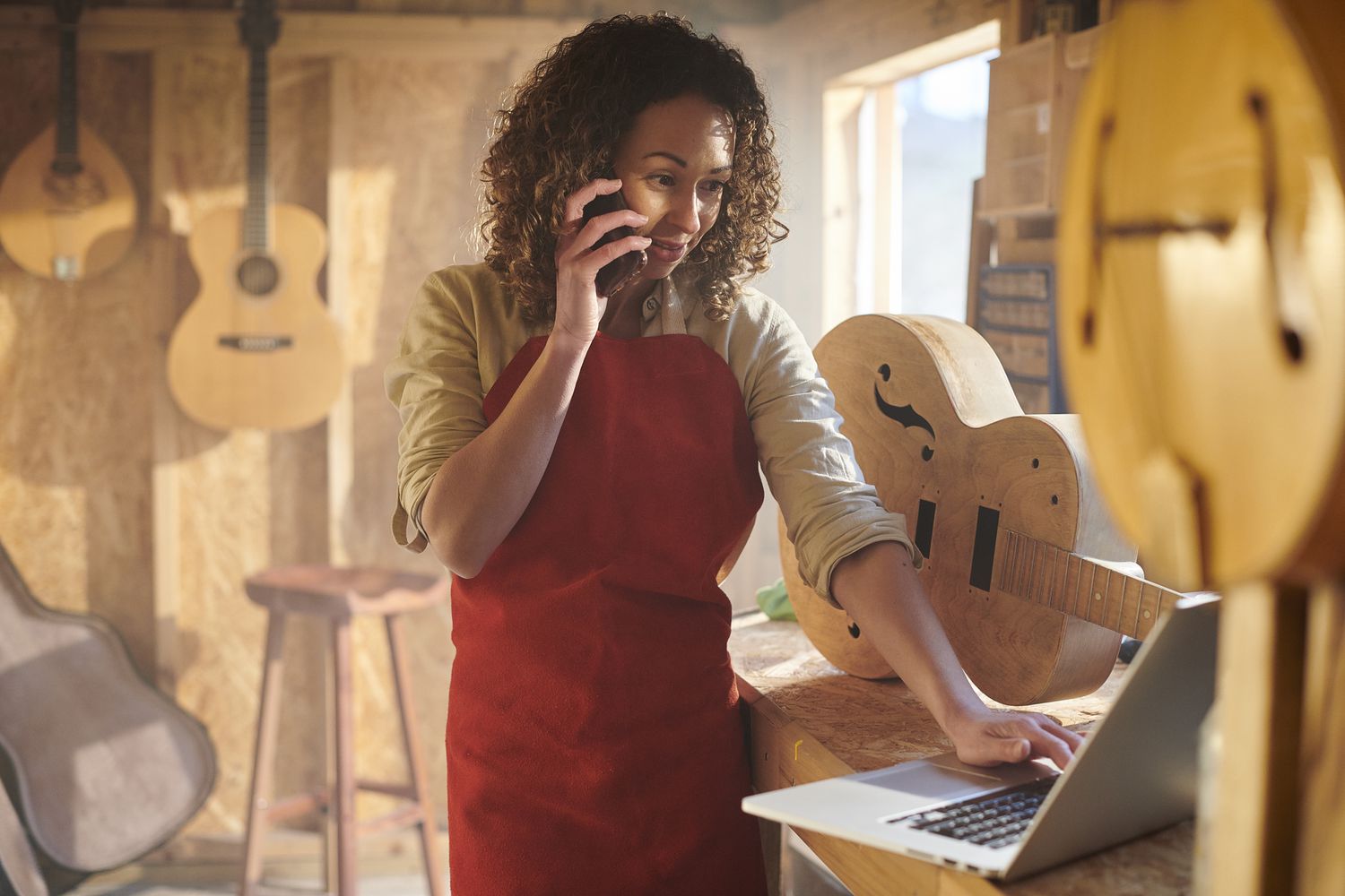 A woman in a guitar shop talks on the phone while reviewing something on a laptop screen