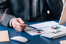 A man wearing a suit sits at a desk and points with a pen to a paper with financial charts.