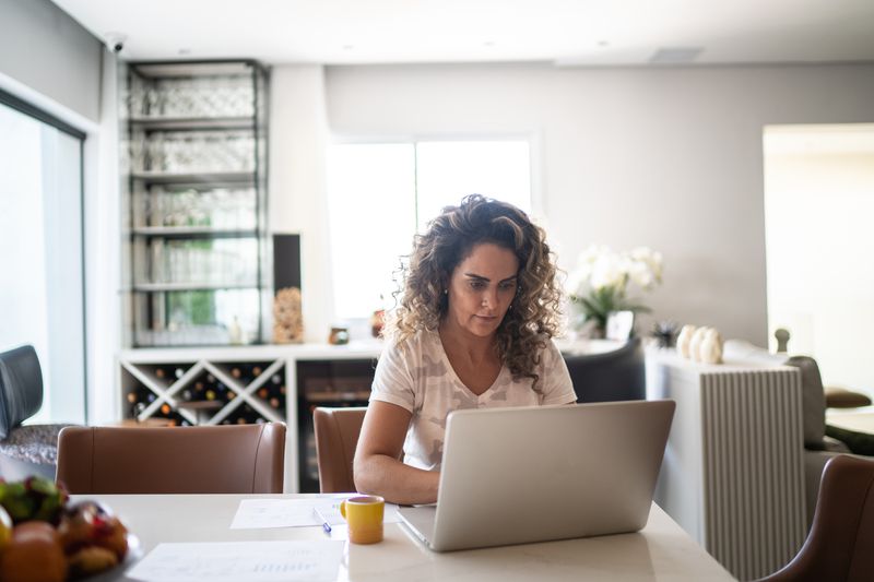 A woman sits at a table in a house with a laptop and small yellow mug