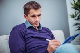 A man sitting on a couch writes something on paper with a pen.