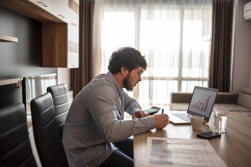 Man writing while sitting at a desk, open laptope displaying data.