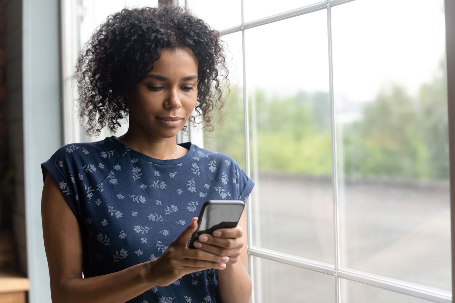 Young woman standing next to a window in her home and looking seriously at something on her smartphone