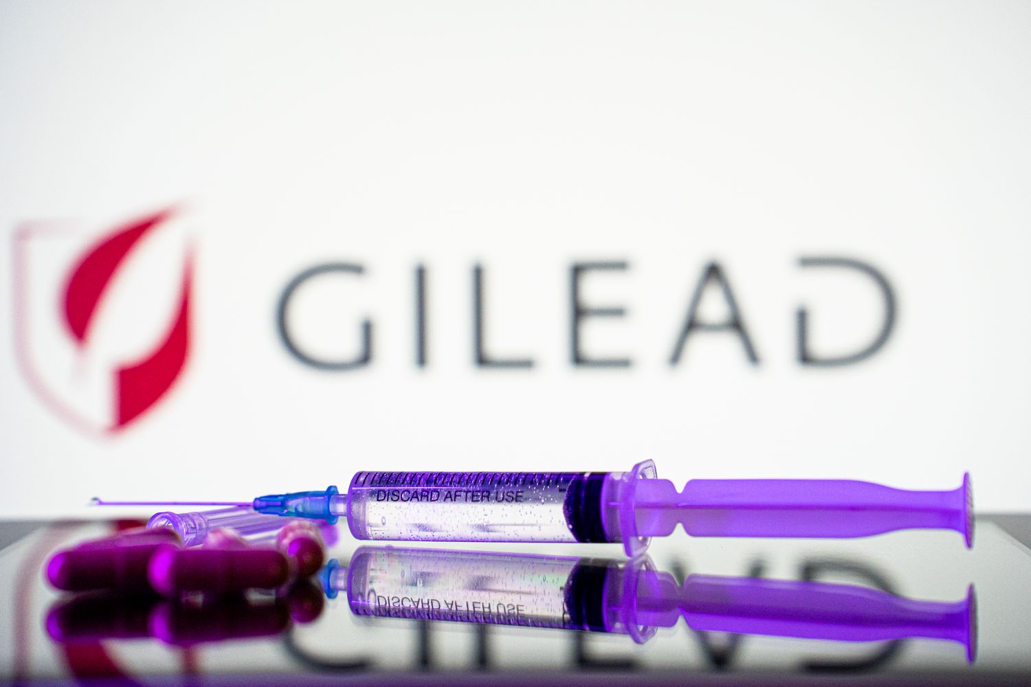 A syringe and some pills are in front of the logo of Gilead pharmaceutical company