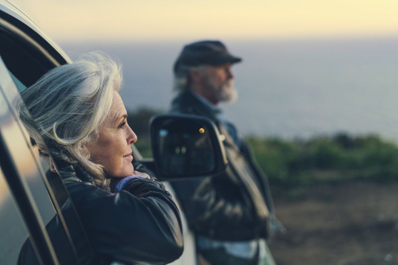 An older woman leans on a car door facing a sunset, while an older man leans against the ood