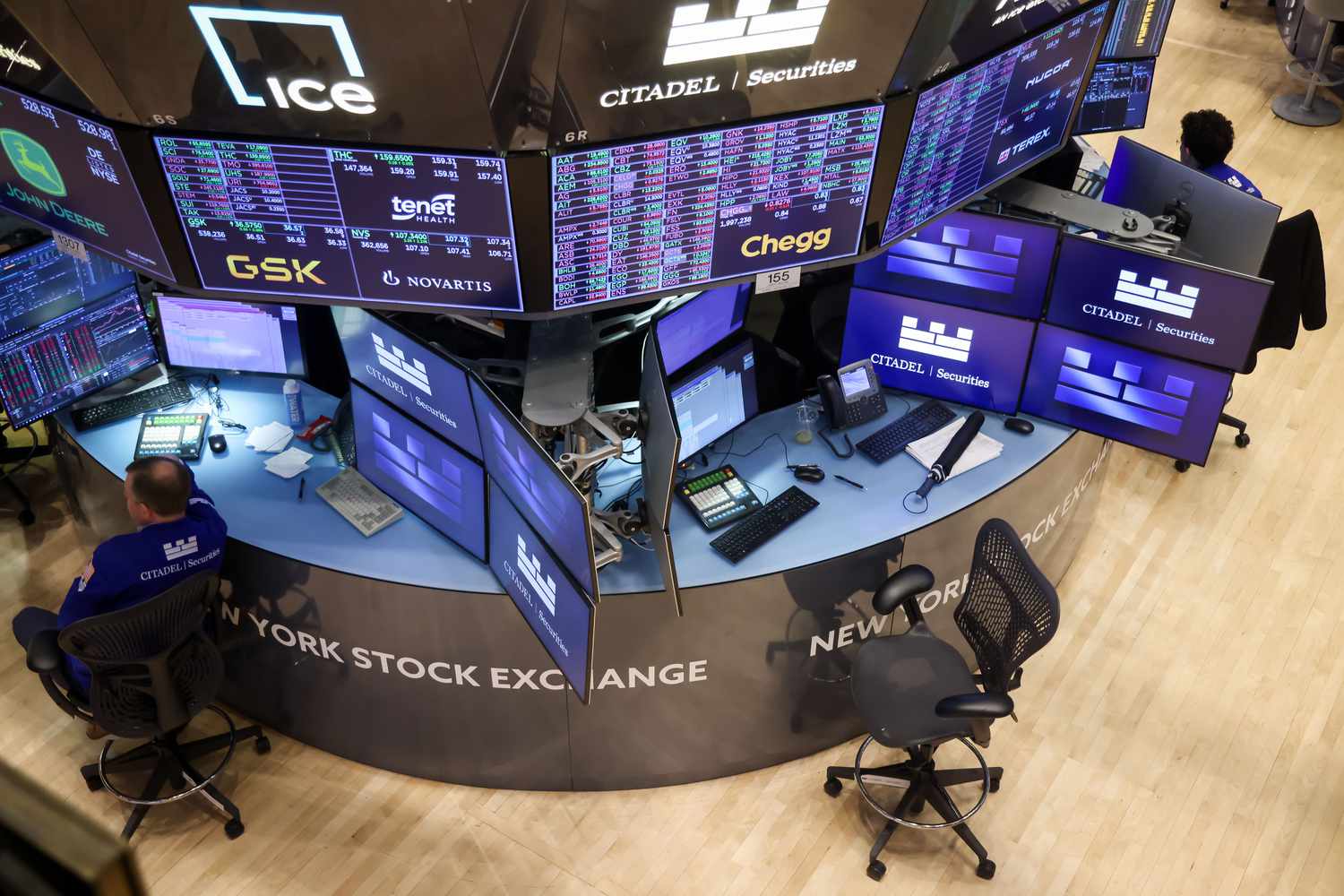 Traders sit at computers on the New York Stock Exchange floor, surrounded by monitors with stock changes and company logos.