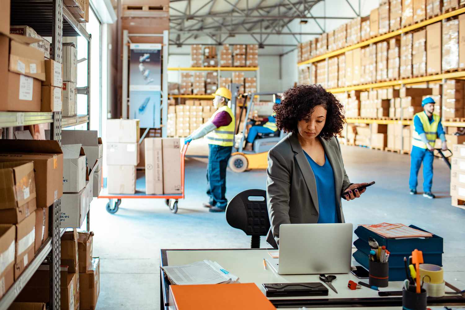 Business manager using laptop in a warehouse with workers moving boxes