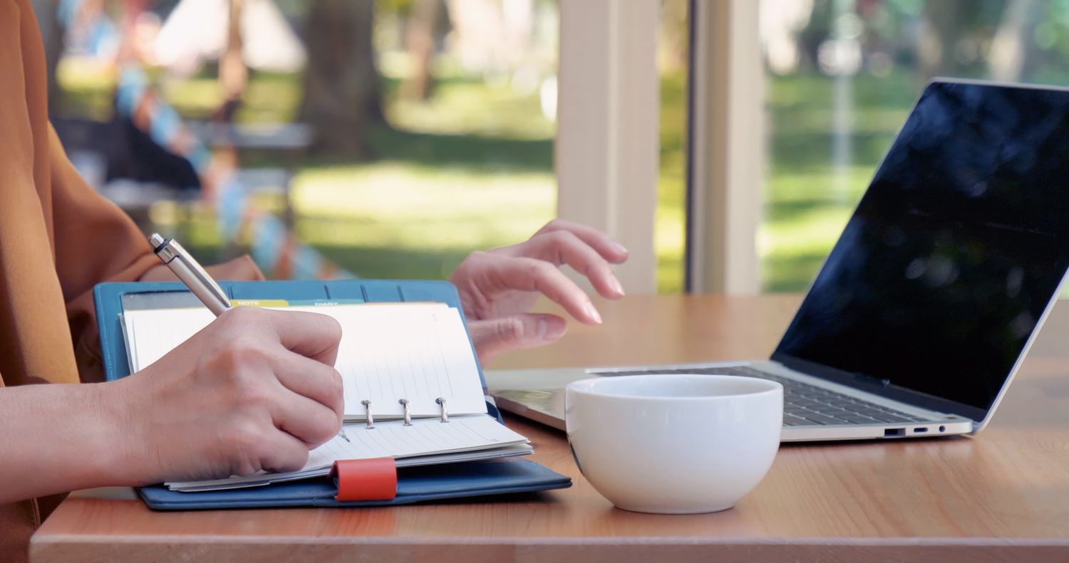 Woman taking notes in a notepad while working on a laptop.
