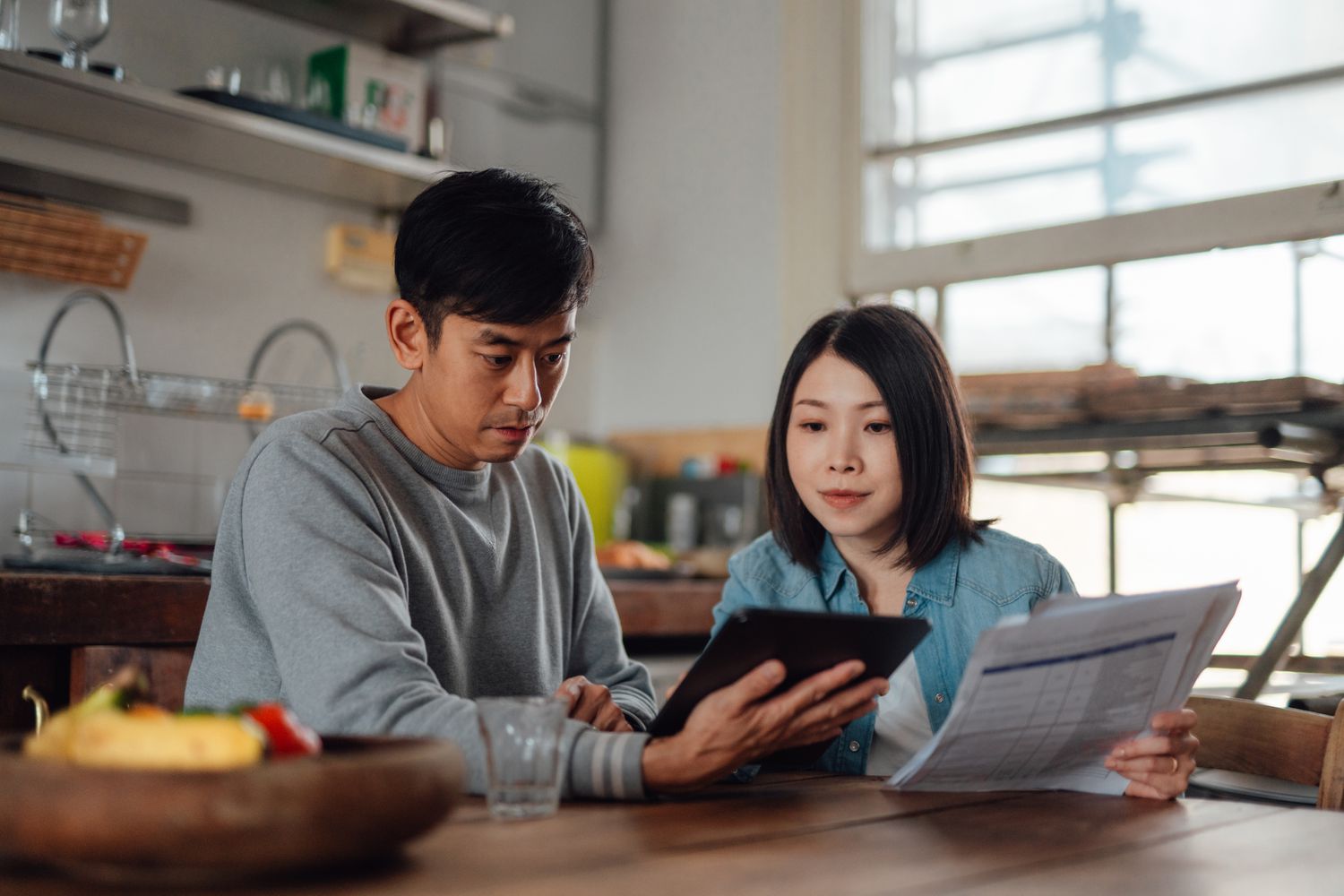 A young couple reviews documents and a tablet computer screen while seated in a sleek, spare, home kitchen.