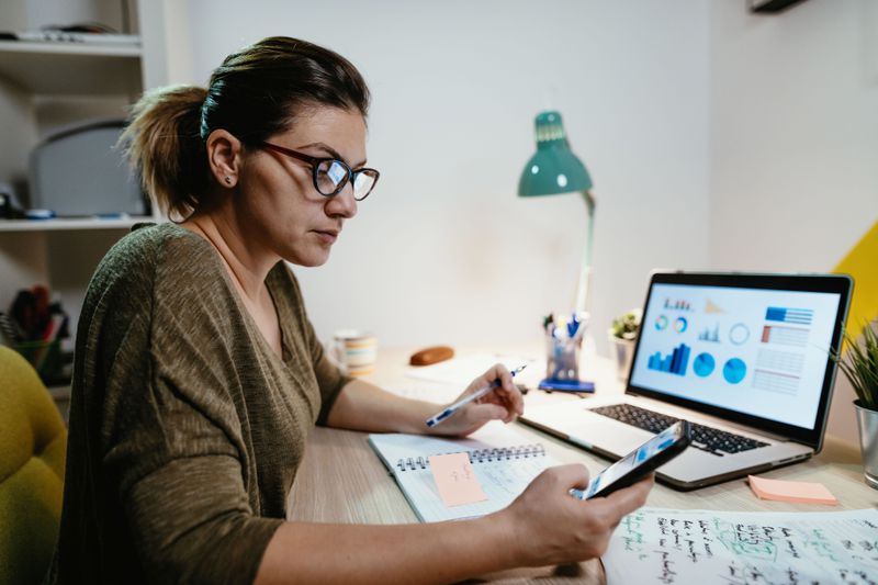 Woman looking at market charts on her smartphone and a laptop, seated at a desk
